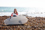 Kate Richards in a white, orange, and black top sits on a beach covered in seaweed, holding a white surfboard with black Rad logos. The ocean is visible in the background under a bright sky.