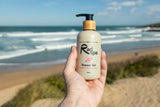 A hand holds a bottle of Rad spa Shower Gel in front of a blurred beach background with ocean waves and sand.