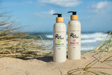 Two Rad Spa Shower Gel 500ml  bottles standing on sand in front of a beach with ocean waves and dune grass in the background.